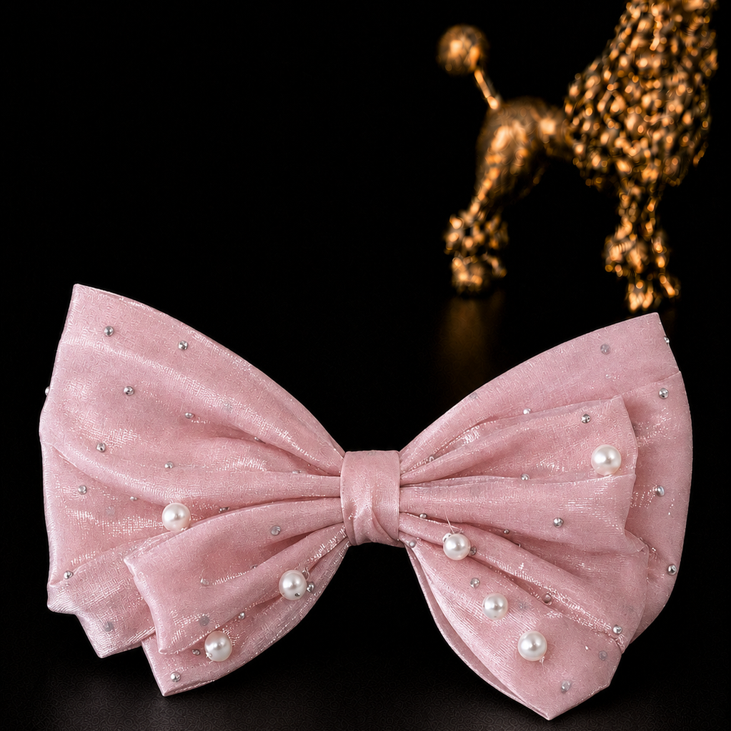 A light pink velvet bow tie decorated with small silver rhinestones and white faux pearls, displayed on a black background with a gold poodle figurine in the background.