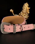 A pink crocodile-embossed leather belt with a large silver-tone buckle embellished with clear rhinestones, displayed on a dark surface with a gold poodle figurine in the background.