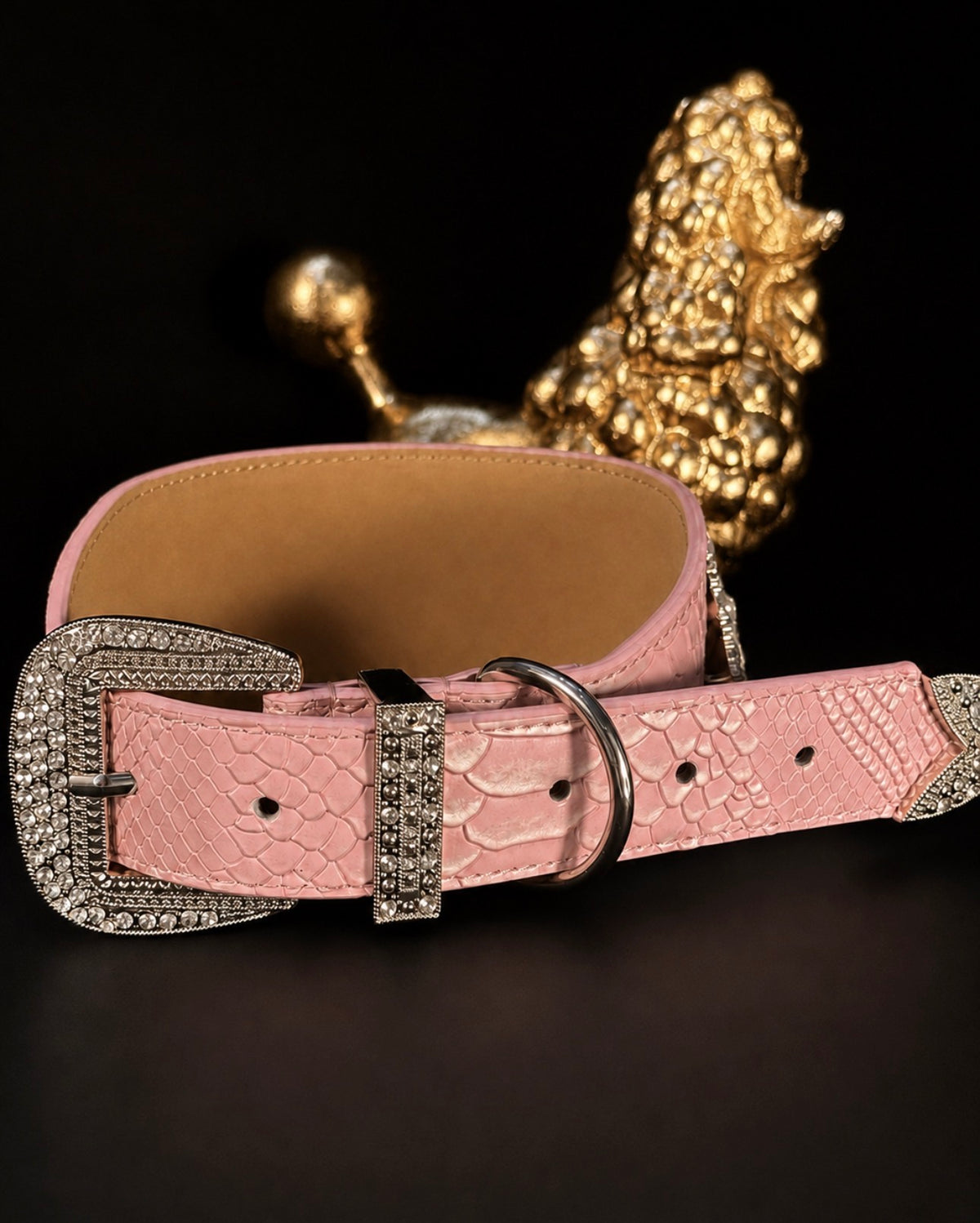 A pink crocodile-embossed leather belt with a large silver-tone buckle embellished with clear rhinestones, displayed on a dark surface with a gold poodle figurine in the background.