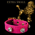 A bright pink faux-leather dog collar with gold-tone hardware and three clear rhinestone studs, shown against a black background with a blurred gold poodle statue and the text 'EXTRA SMALL' at the top.